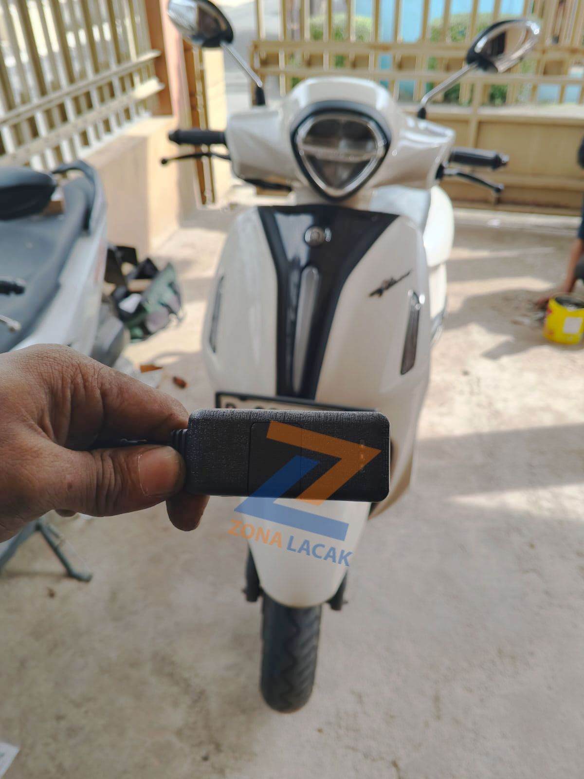 Perangkat GPS Tracker berlogo Zona Lacak dipegang di depan sepeda motor skuter
